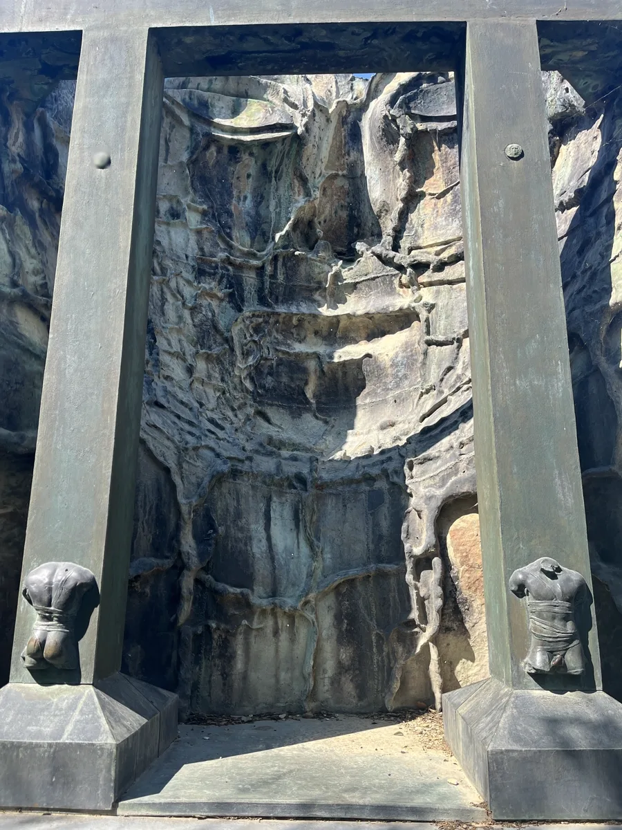 Wide detail of a monumental Mitoraj bronze relief flanked by two vertical piers with small torso sculptures at each base, deep chiaroscuro across the fractured surface