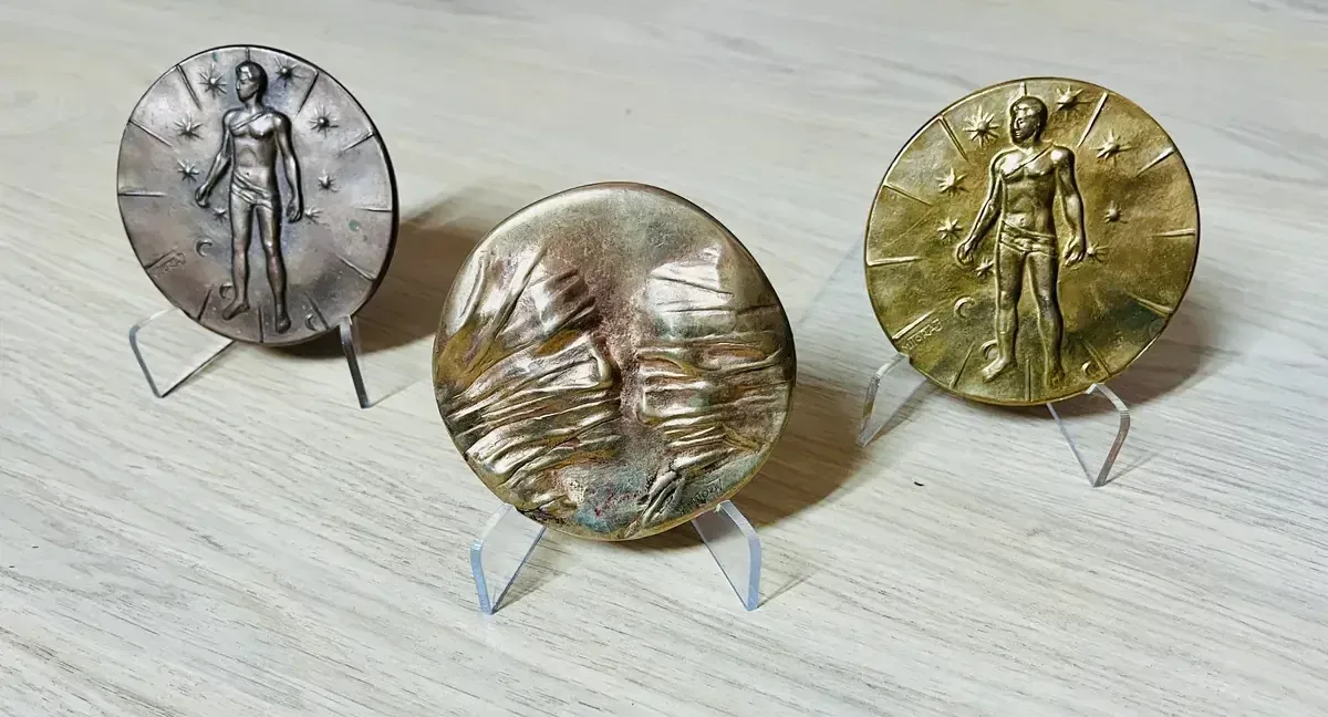 La Conversation — Igor Mitoraj, three patina variants on acrylic stands: dark reverse, iridescent obverse, gilt reverse, wood surface