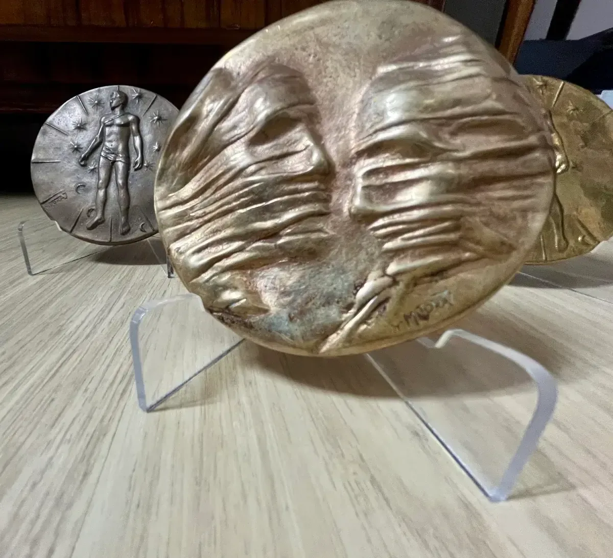 La Conversation — Igor Mitoraj, three specimens low-angle on granite: gilt reverse, worn obverse, dark reverse, acrylic stands — gold-patina bronze medal
