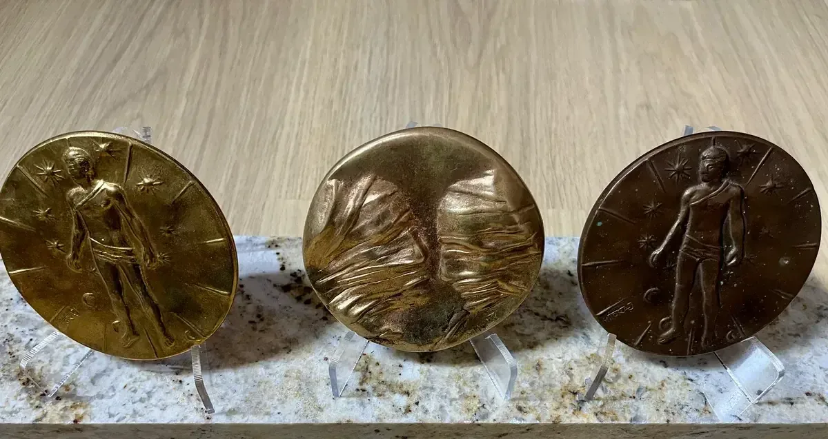 La Conversation — Igor Mitoraj, three specimens on granite: gilt reverse (left), worn obverse (centre), dark brown reverse (right) — gold-patina bronze medal