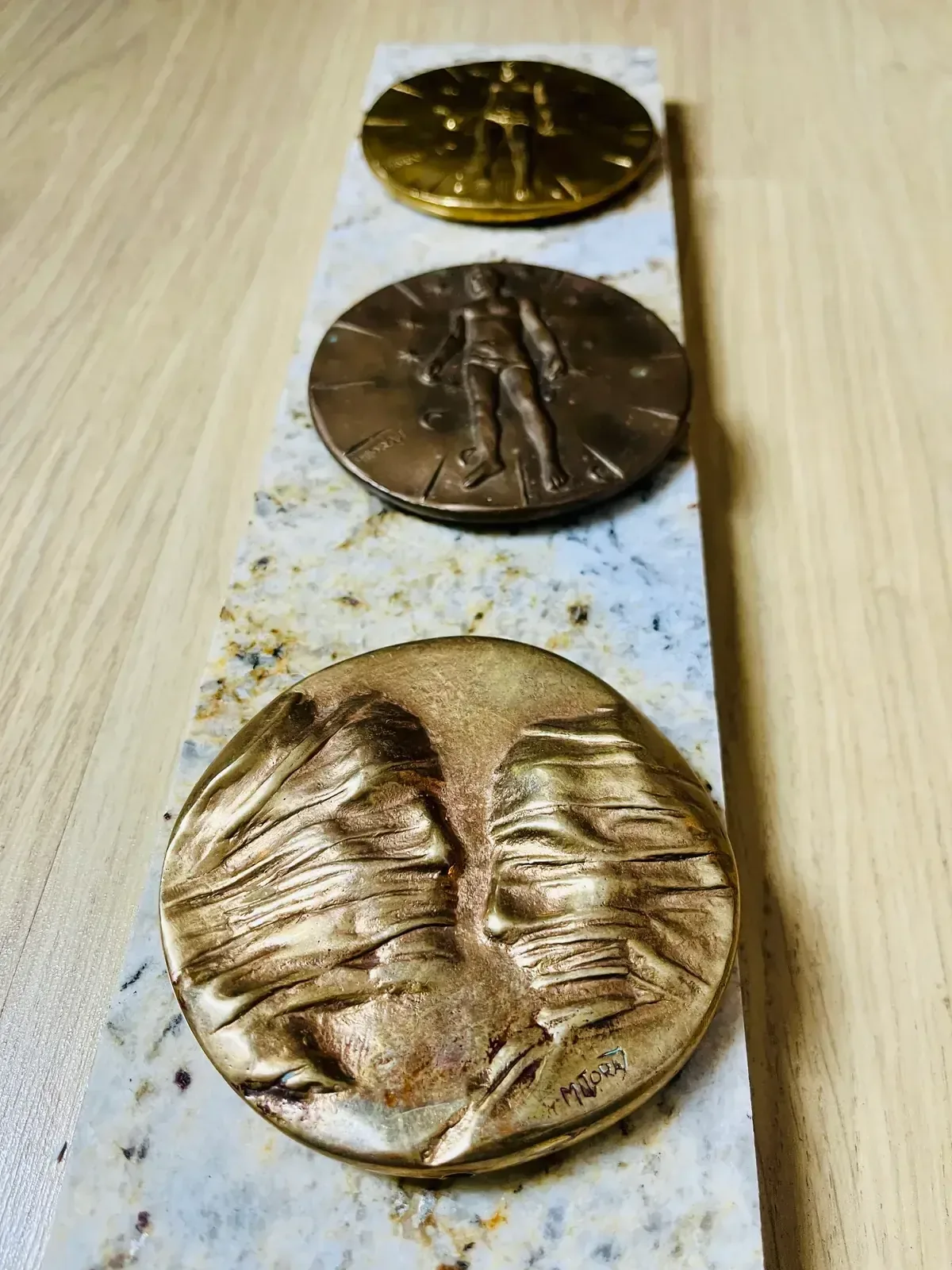 La Conversation — Igor Mitoraj, three medals vertical on marble: gilt reverse (top), dark reverse (middle), worn obverse (bottom)