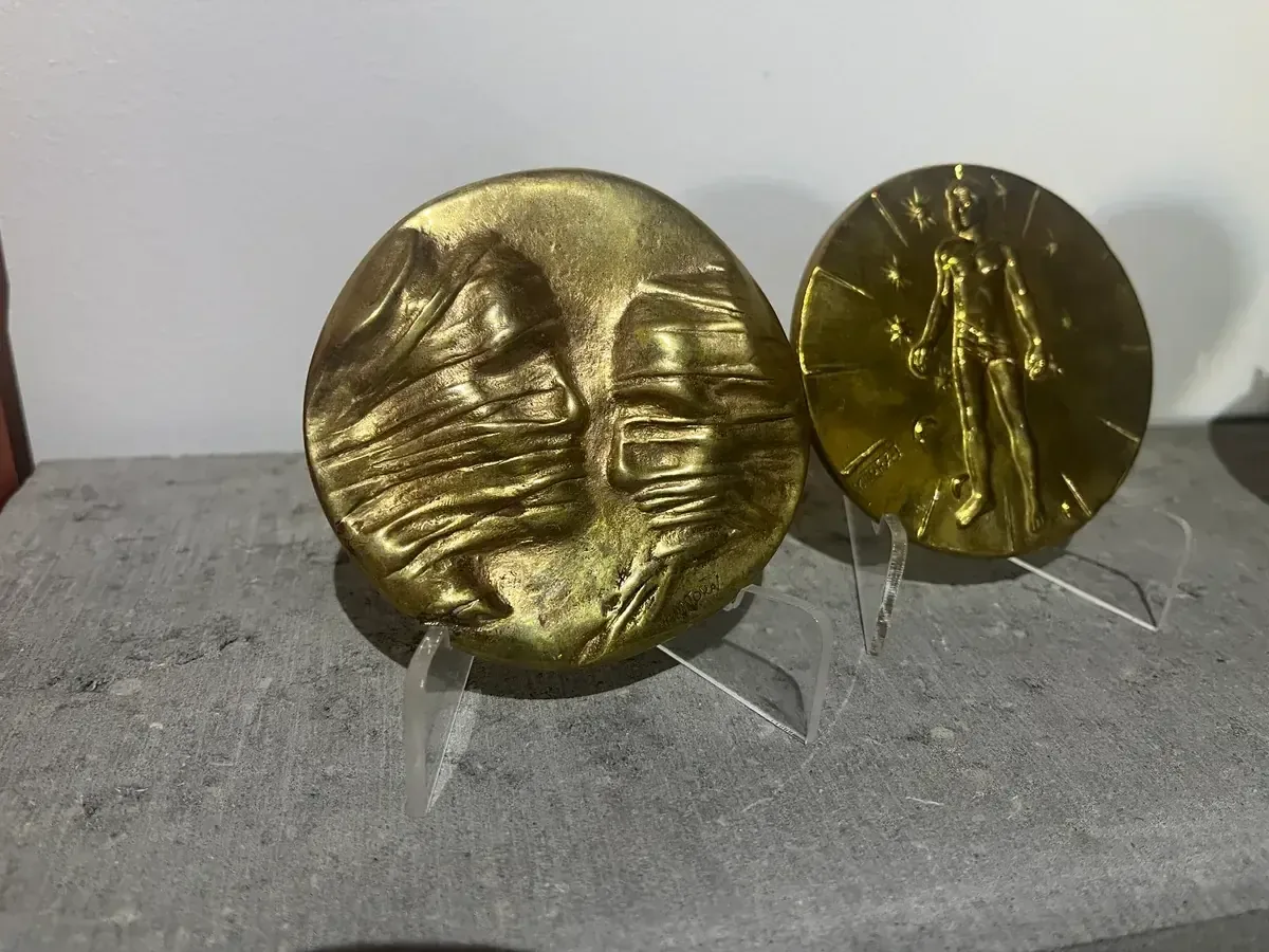 La Conversation — Igor Mitoraj, both medal variants on acrylic stands, obverse and reverse side by side — gold-patina bronze medal