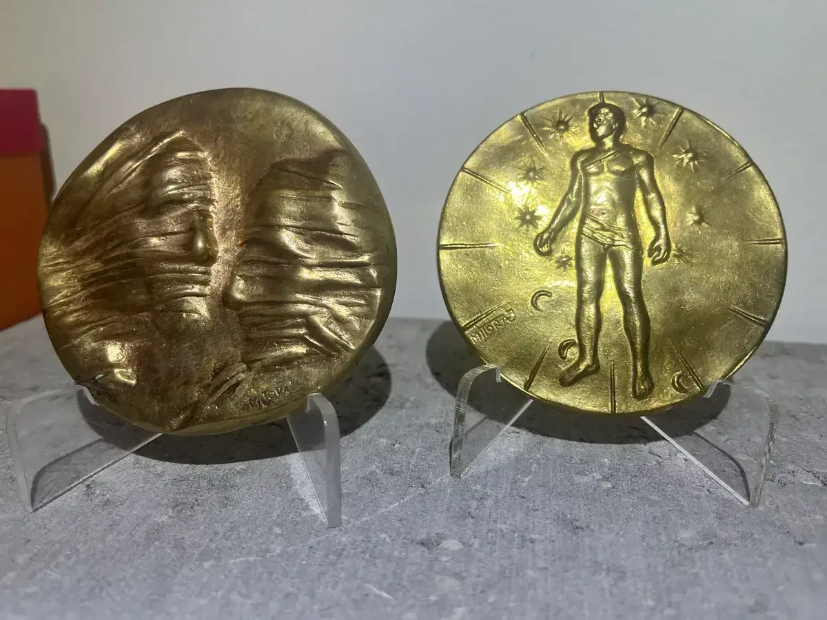 La Conversation — Igor Mitoraj, both medals on stands — obverse showing conversation relief, reverse with standing figure — gold-patina bronze medal