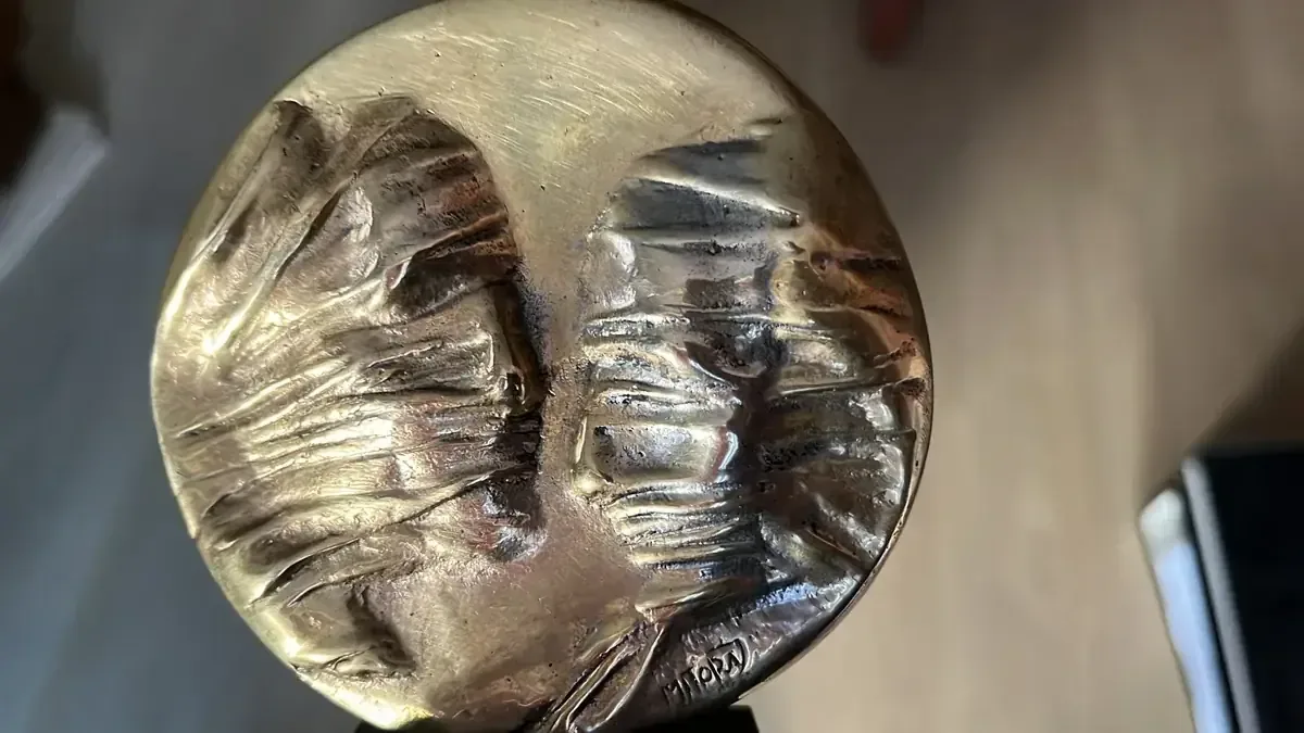 La Conversation — Igor Mitoraj, close-up of obverse: two veiled faces meeting at centre, MITORAJ signature visible, warm gold patina